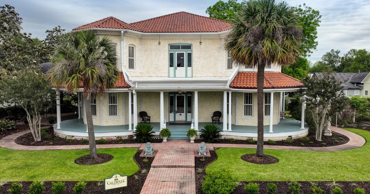 Front elevation of The Caldwell House, a historic estate with a red tile roof, wraparound porch, and palm-lined brick walk
