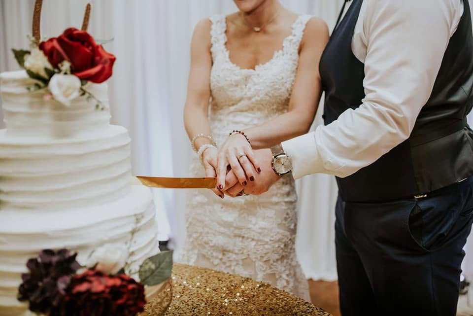 A bride and groom cutting a tiered white wedding cake trimmed with red roses on a gold sequin table, at a reception served by The Southern Spread