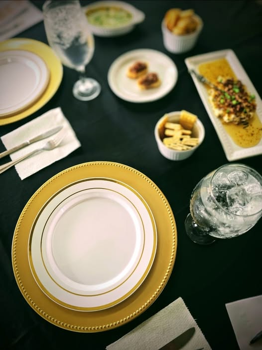 An elegant gold-rimmed place setting on black linen, surrounded by hors d'oeuvres prepared by The Southern Spread